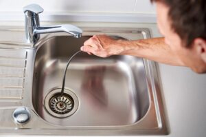 A person is using a plumber's snake tool to clear a clog from a stainless steel kitchen sink.