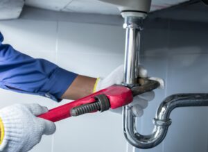 Person wearing gloves using a red pipe wrench to tighten or loosen a metal pipe under a sink.