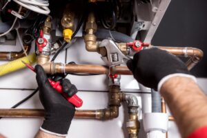 A person wearing gloves uses a wrench and screwdriver to adjust copper pipes and valves connected to a boiler or heating system.