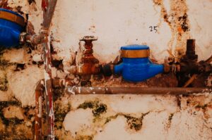 %Title% | Pf Plumbing A blue water meter and rusty valve are attached to old, stained pipes against a weathered, peeling white wall marked with the number 25. Winter Pipe Prep