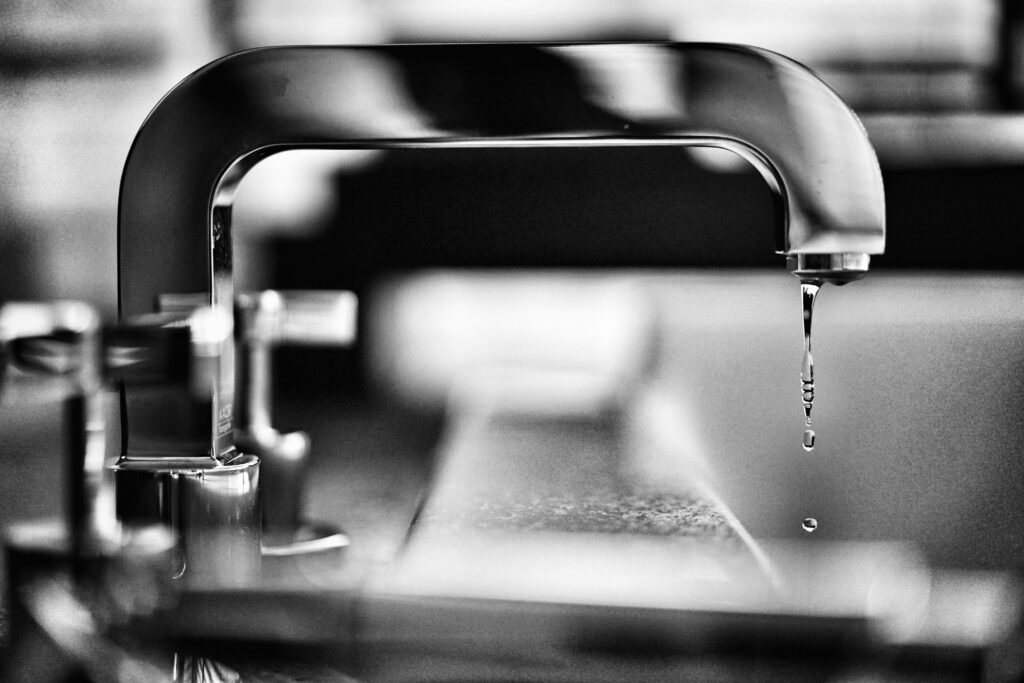 A close-up of a metal faucet with water dripping into a sink. The image is in black and white, with a blurred background.