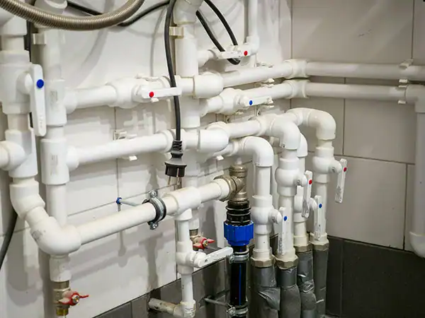Complex arrangement of white PVC water pipes, valves, and connectors mounted on a tiled wall, part of a plumbing or heating system.